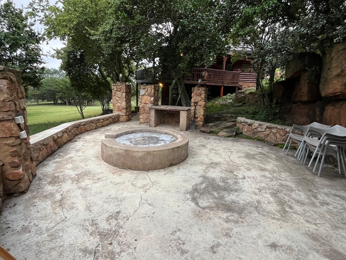 An outdoor fire pit area is surrounded by natural stone walls and trees, creating a tranquil space. Several gray folding chairs are neatly arranged nearby. The ground is made of concrete, with a rustic feel complemented by the nearby stone structure.