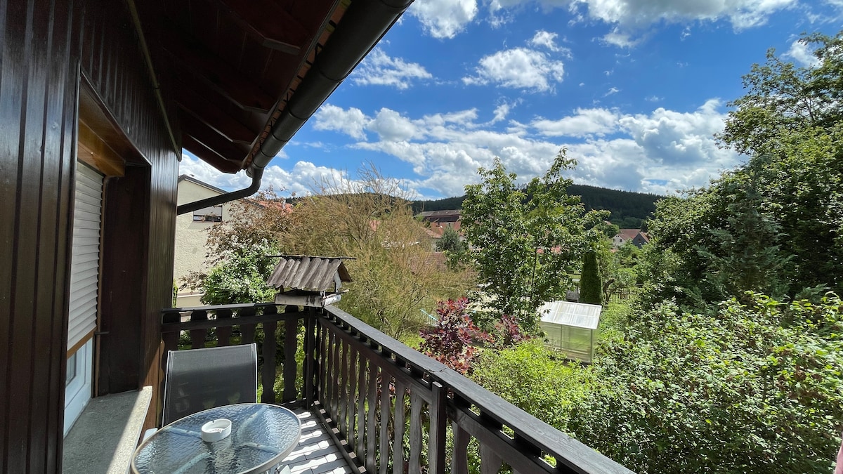 A private balcony is featured, surrounded by greenery and trees. A small glass table with two chairs invites relaxation. In the background, a view of distant hills and a clear blue sky dotted with clouds is visible, creating a serene outdoor atmosphere.