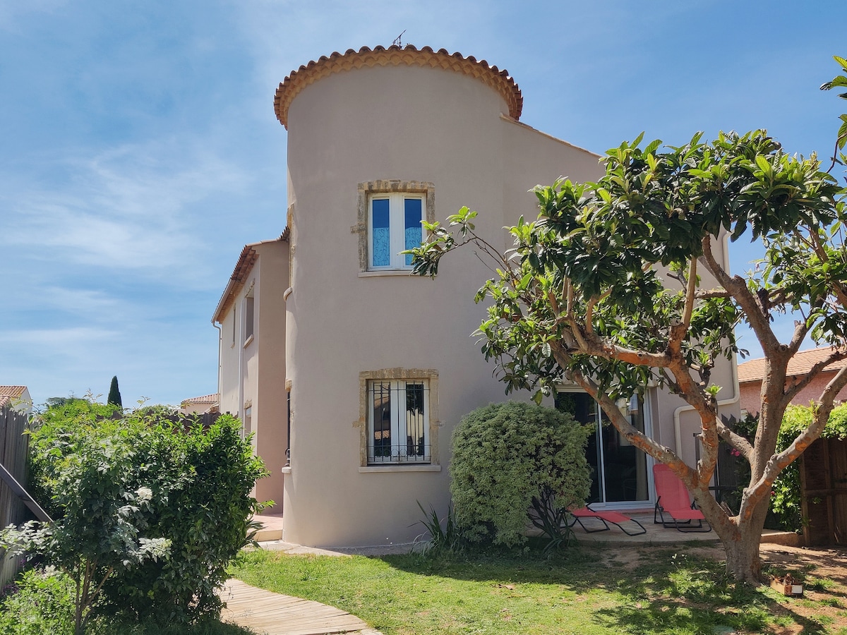 The exterior of the villa is showcased, featuring a round turret and a charming garden with lush greenery. A wooden pathway leads to the entrance, while sun loungers are visible on the terrace. The building has several windows allowing natural light to brighten the space.