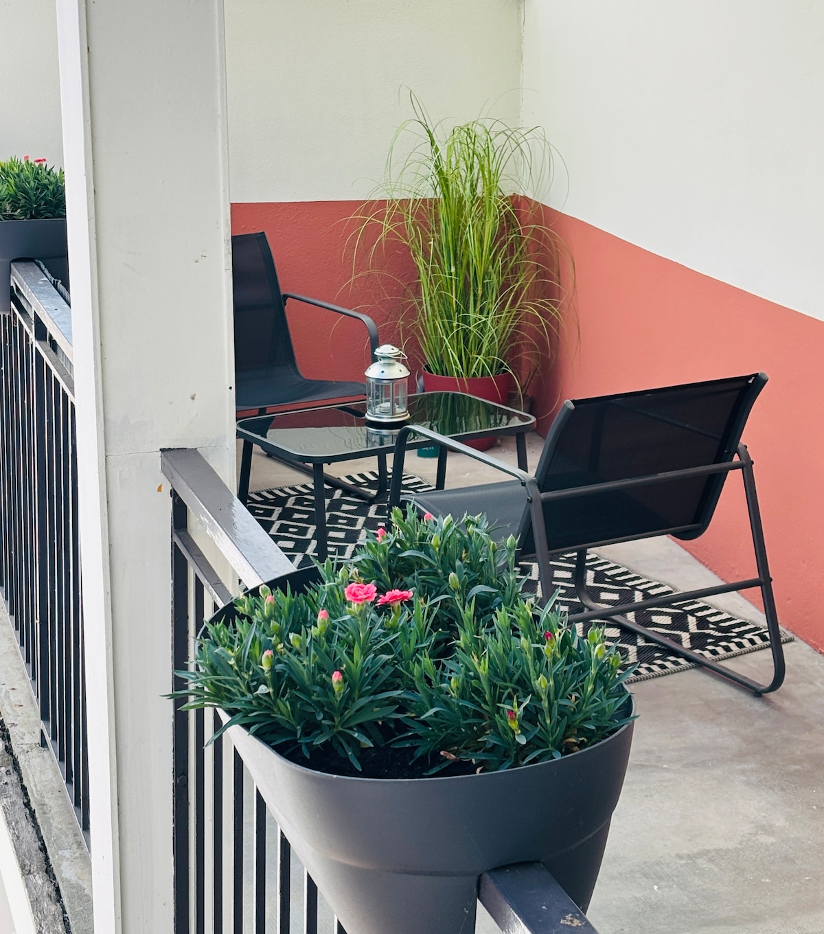 A private terrace features a small table and two chairs, surrounded by decorative plants and greenery. A patterned rug adds texture to the space, while a lantern sits on the table, complementing the relaxing setting.