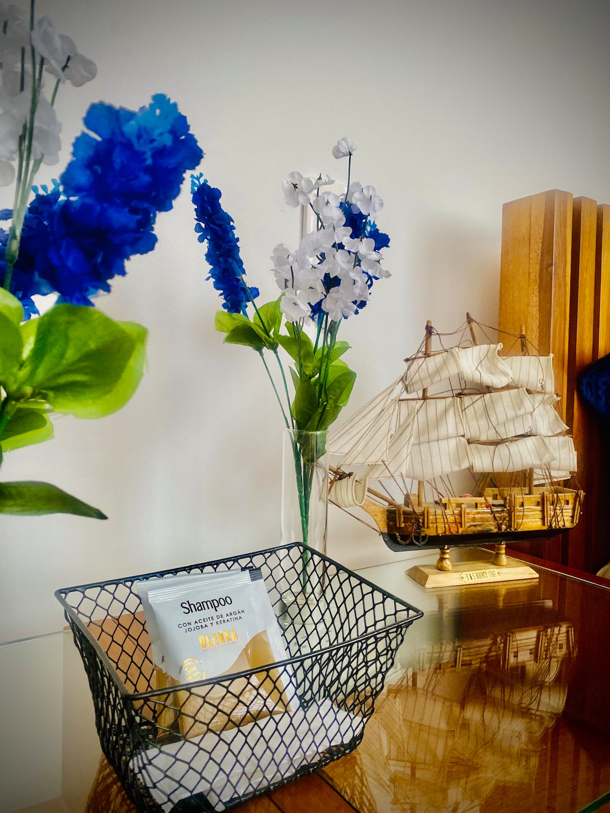 A decorative arrangement features a black wire basket containing shampoo on a polished surface. Bright blue and white faux flowers are placed in a clear vase nearby. A detailed model ship provides an artistic touch, complementing the serene ambiance of the space.