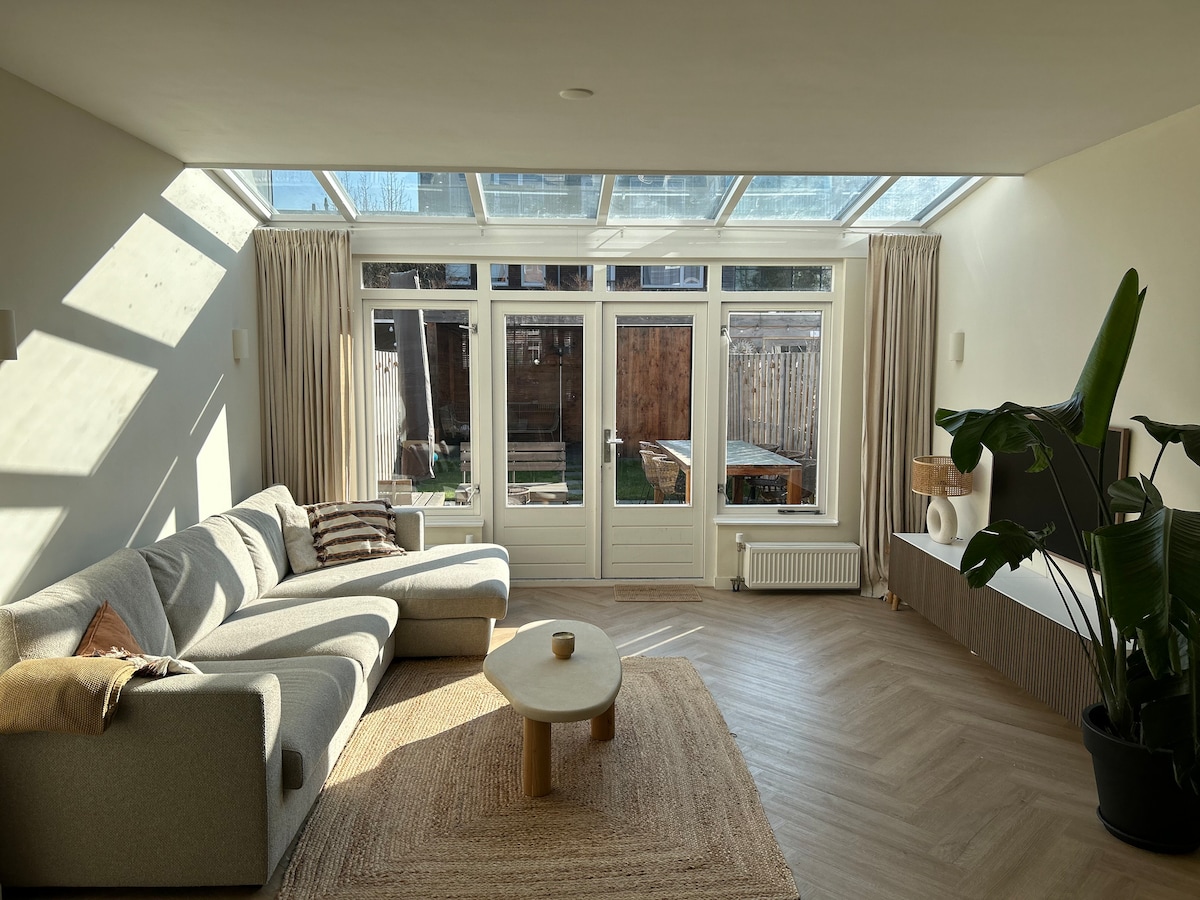 A spacious living area filled with natural light from large glass panels above. A comfortable sectional sofa is arranged near a light-colored coffee table, with a woven rug beneath. Sliding doors lead to an outdoor garden space, offering a seamless connection to nature.