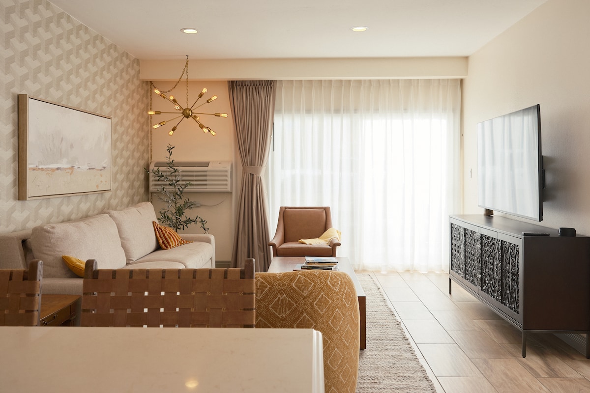 A spacious living area features a light-toned sofa paired with a stylish armchair. A large TV is mounted on a dark wooden console, and natural light filters in through sheer curtains. Decorative touches include a modern light fixture and wall art, adding warmth to the space.