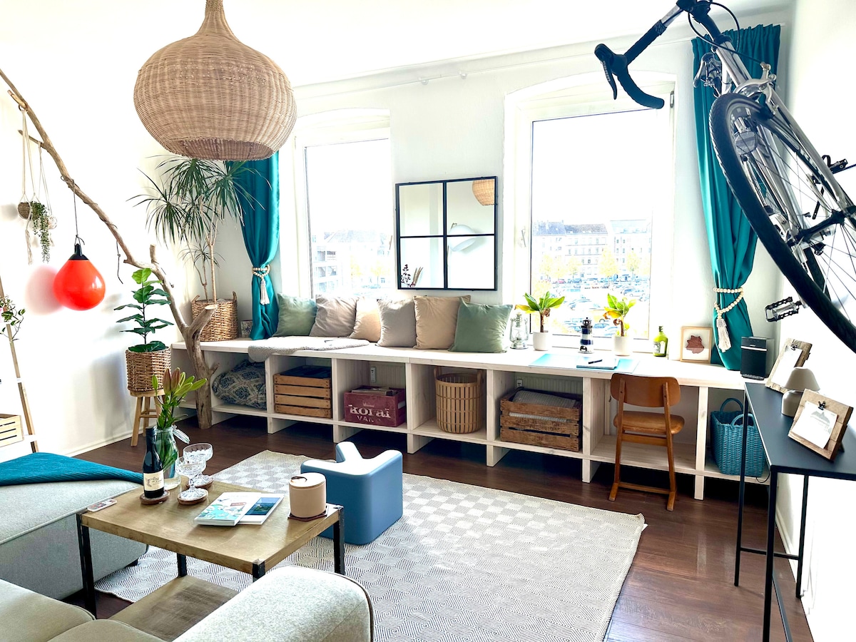A sunlit living area features large windows adorned with teal curtains, offering a view of the surroundings. A cozy couch invites relaxation beside a wooden coffee table. Decorative elements include a bicycle mounted on the wall and potted plants enhancing the space's welcoming feel.