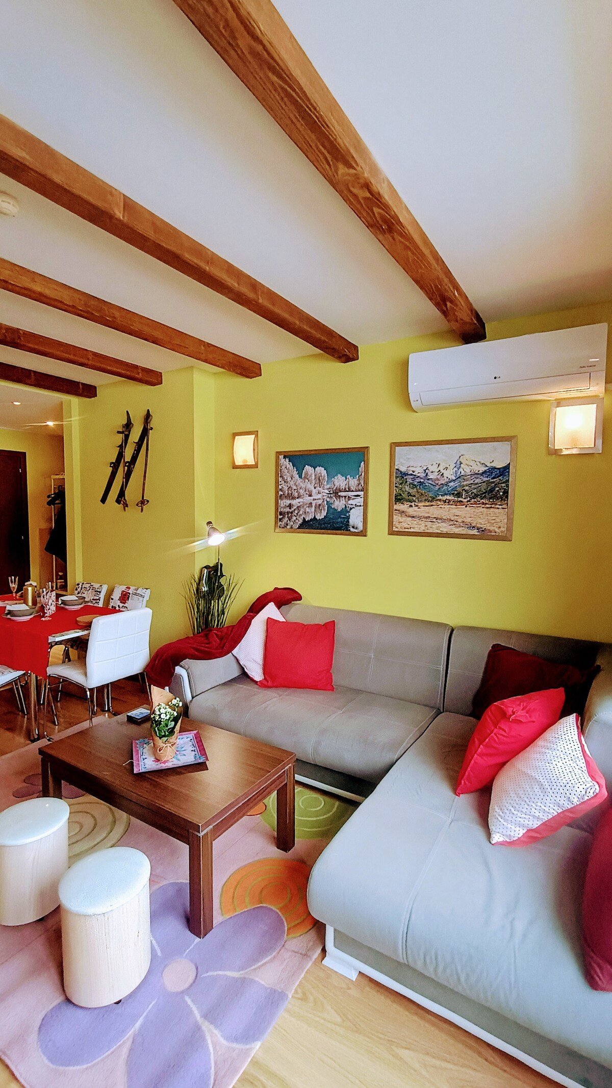 A cozy living area features a comfortable sectional sofa adorned with red cushions, complemented by a wooden coffee table. Colorful wall art enhances the atmosphere, while ski equipment decorates the space. Warm lighting and natural tones create an inviting environment for relaxation.
