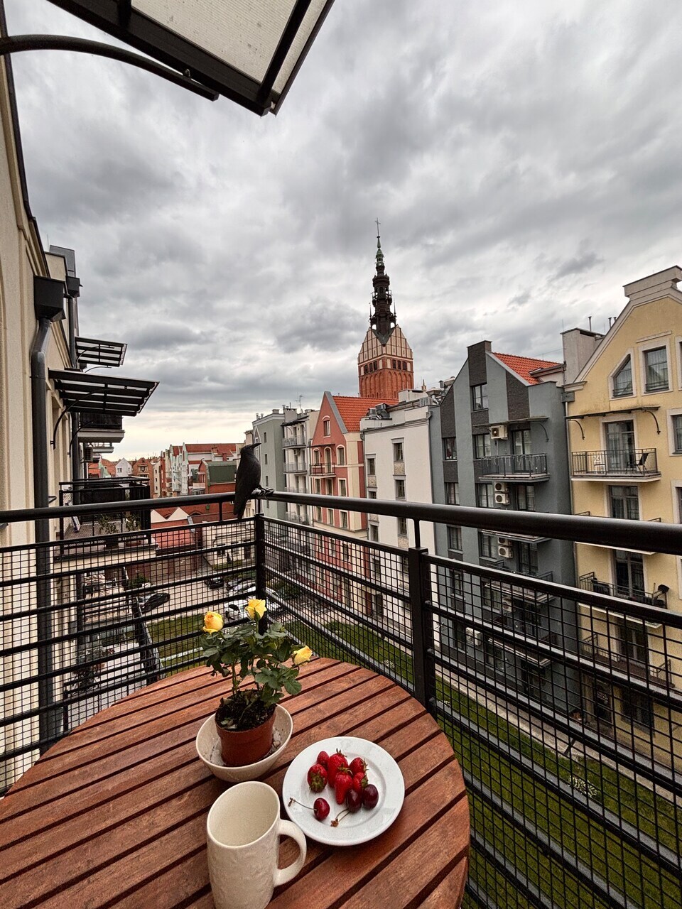 A balcony table made of wood features a small potted plant and a plate of strawberries. Surrounding buildings are visible, along with a prominent tower in the distance under a cloudy sky. A railing provides a view of the neighborhood below.