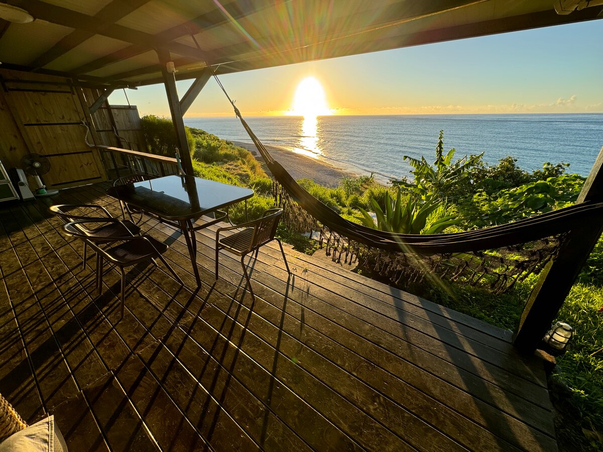 A spacious deck features a glass table and four chairs, surrounded by lush greenery. A hammock is draped nearby, offering a relaxing spot. The view includes a stunning sunset over the water, creating captivating reflections on the surface.