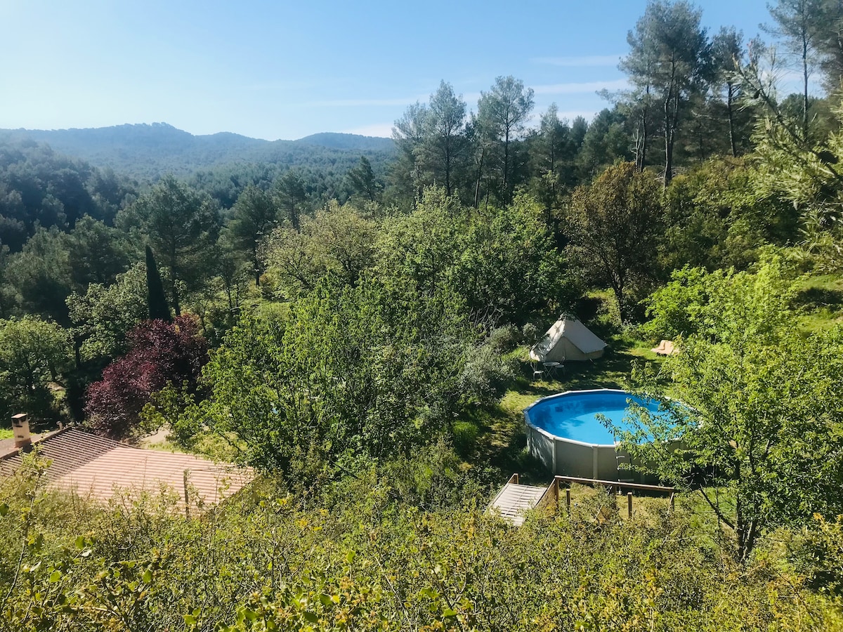 A spacious outdoor setting is captured, featuring a round above-ground pool surrounded by trees and greenery. In the distance, a glamping tent is visible, nestled among the trees. The natural landscape offers a serene view of rolling hills and clear skies.