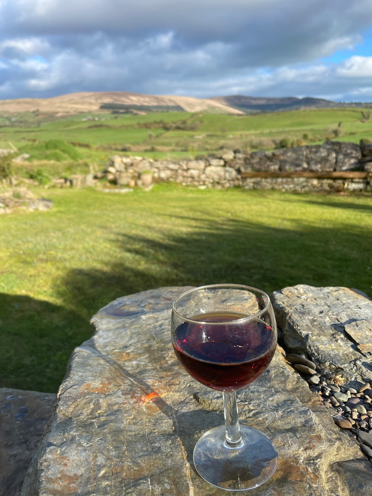 A glass of red wine is placed on a stone table, with a lush green landscape in the background. Rolling hills and an expansive sky can be seen, reflecting a serene atmosphere. The scene invites relaxation and enjoyment of nature's beauty.