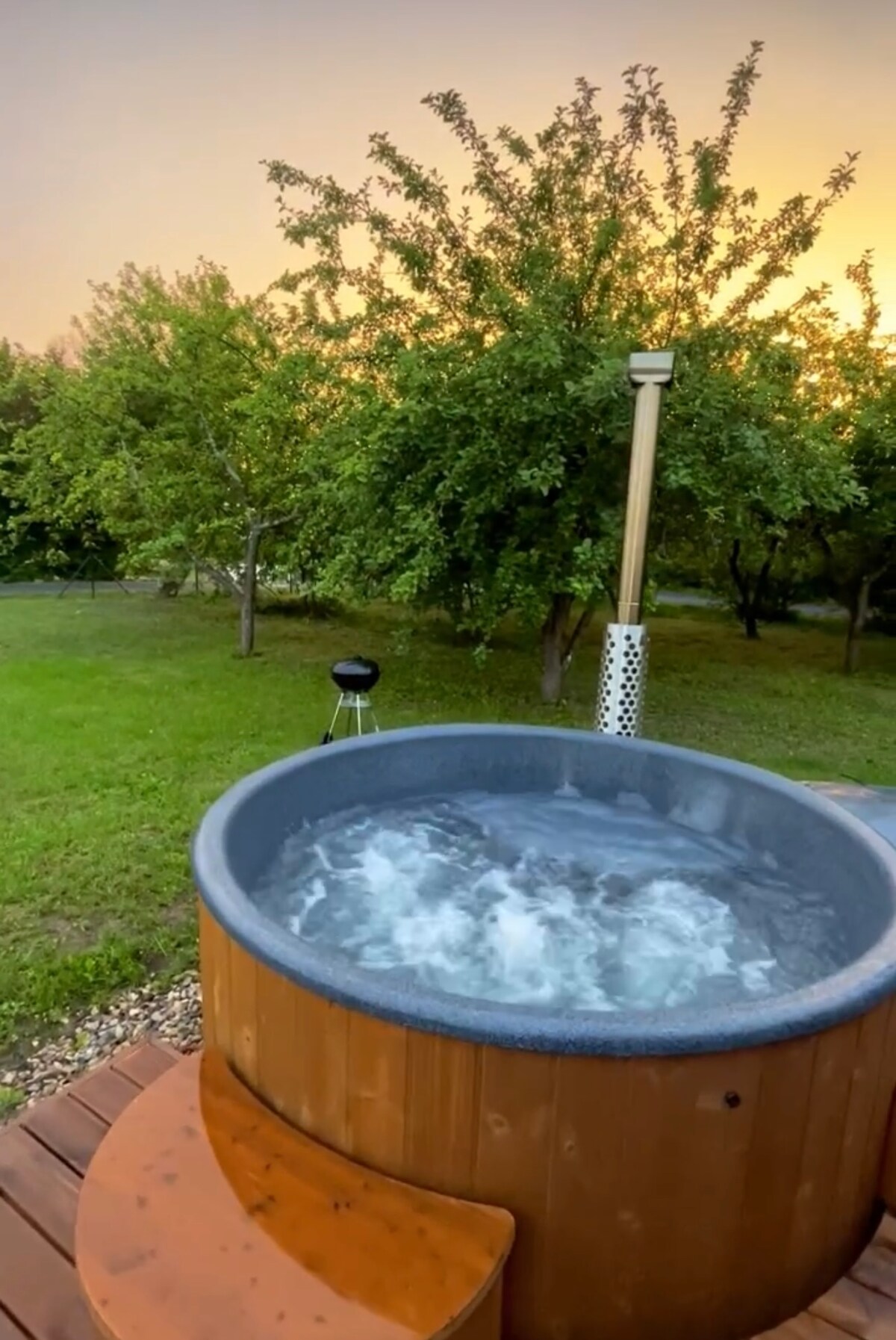 A wooden hot tub filled with bubbling water is positioned on a wooden deck. Lush greenery surrounds the area, with trees providing a natural backdrop. A small grill is visible nearby, enhancing the outdoor setup.