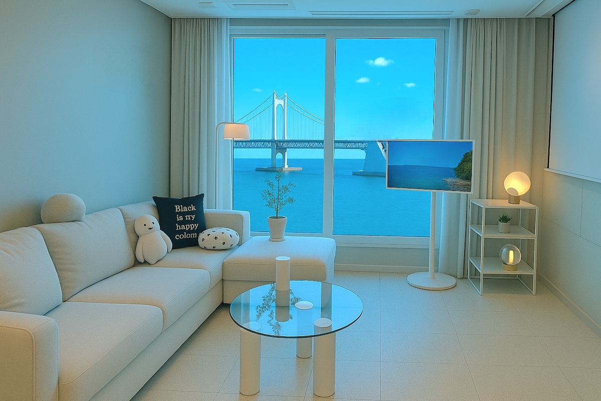 A modern living area features a white sofa complemented by decorative pillows, a glass coffee table, and minimalistic shelving. A large window provides a stunning view of the ocean and Gwangandaegyo Bridge, while a television stands nearby, enhancing the inviting atmosphere.