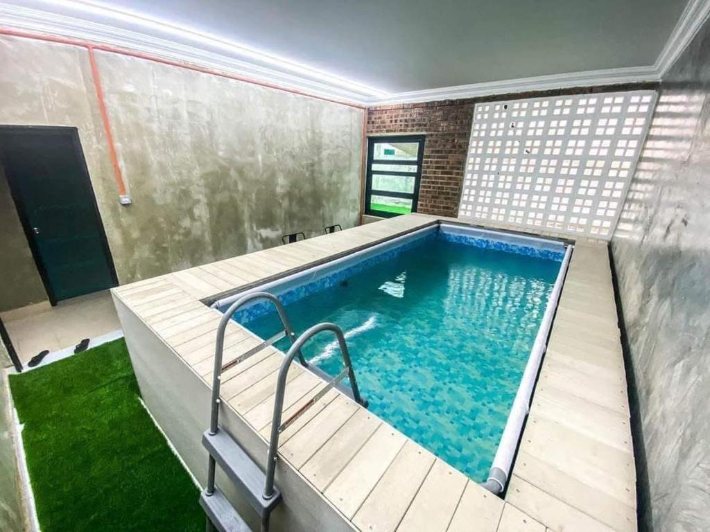 An indoor swimming pool is featured within a modern space, surrounded by textured walls and wooden decking. A metal ladder provides easy access to the water, which reflects soft lighting from the ceiling. Adjacent greenery adds a touch of nature to the serene environment.
