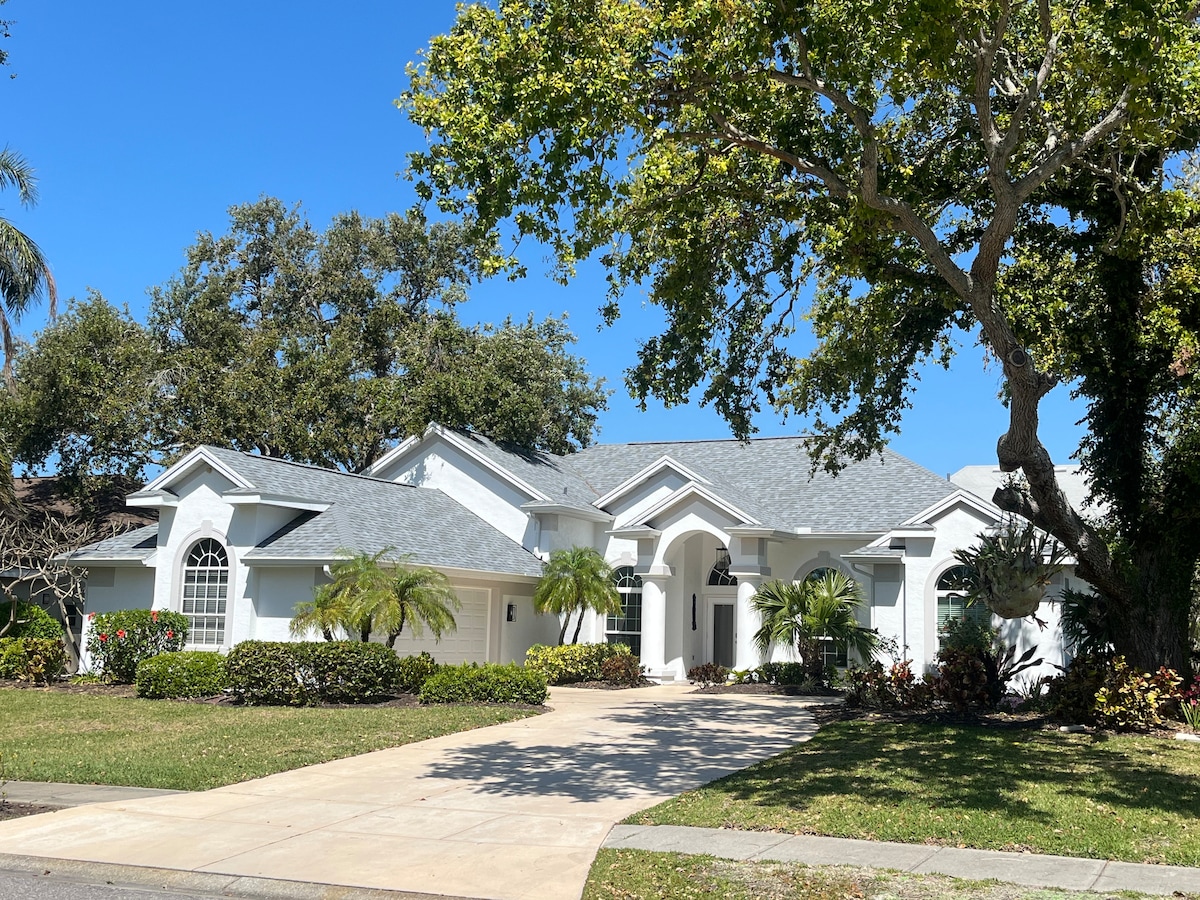 A modern home is depicted, featuring a light exterior with multiple large windows and an inviting entrance. Lush greenery and landscaped gardens frame the property, providing a serene atmosphere. A spacious driveway leads up to the house, complemented by a clear blue sky.