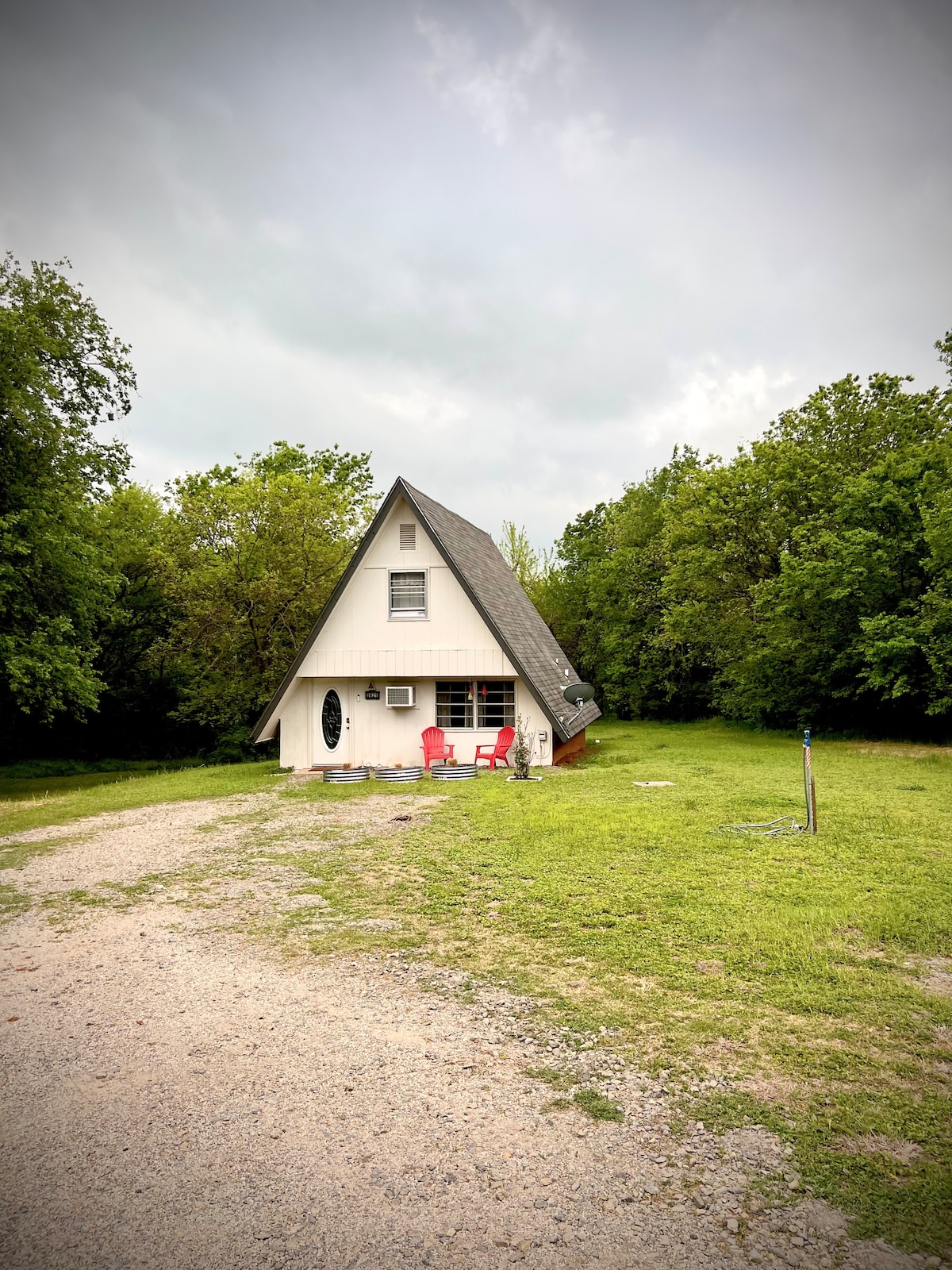 The A-frame home is set in a green landscape, surrounded by trees. A gravel path leads to the entrance, where two red chairs are positioned on the porch. The exterior features a white facade and a sloped roof, offering a welcoming appearance in the countryside.