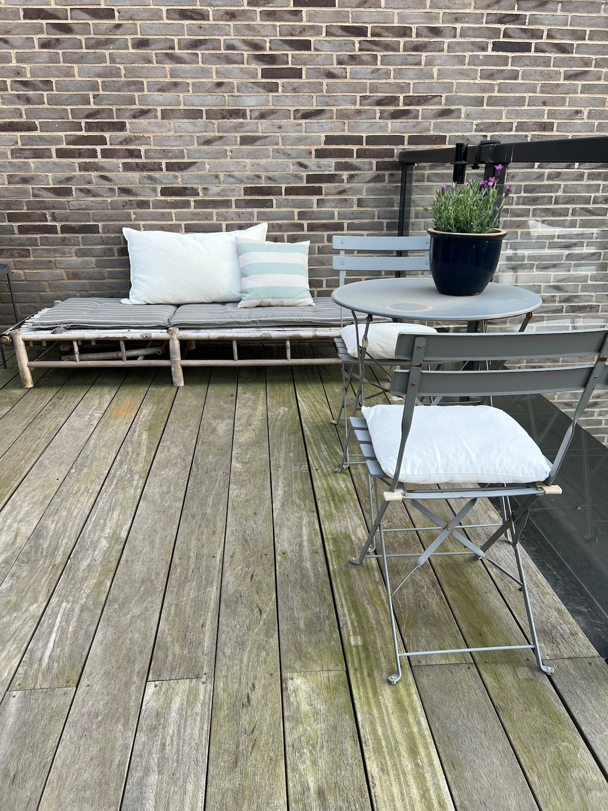 A cozy terrace area features a wooden floor with a small round table and two folding chairs. A potted plant is placed on the table. Alongside, a comfortable lounge area consists of a cushioned bench adorned with decorative pillows.