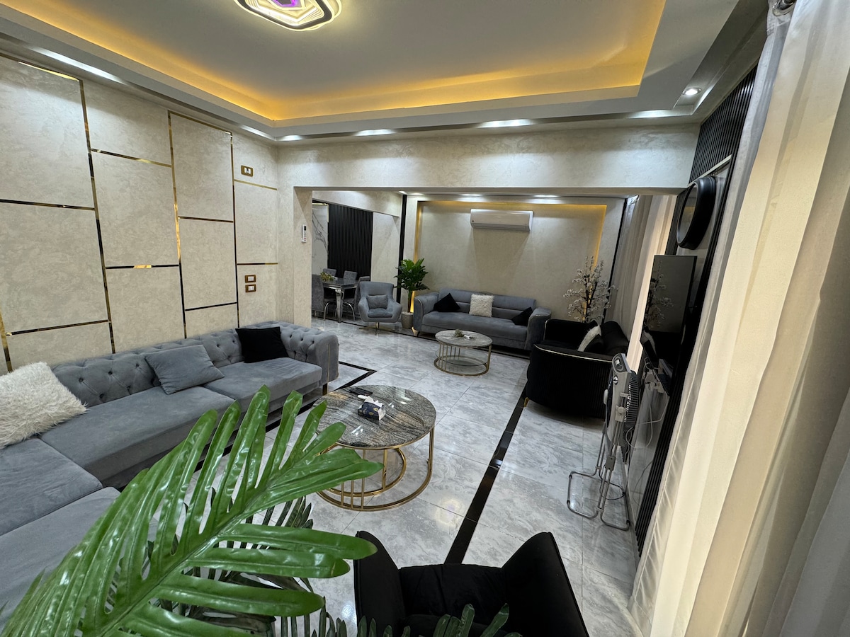 A spacious living room is presented, featuring a combination of plush seating arranged around a circular coffee table. Neutral-colored walls are complemented by subtle lighting, enhancing the modern design. A decorative plant adds an element of greenery, contributing to a relaxed atmosphere.