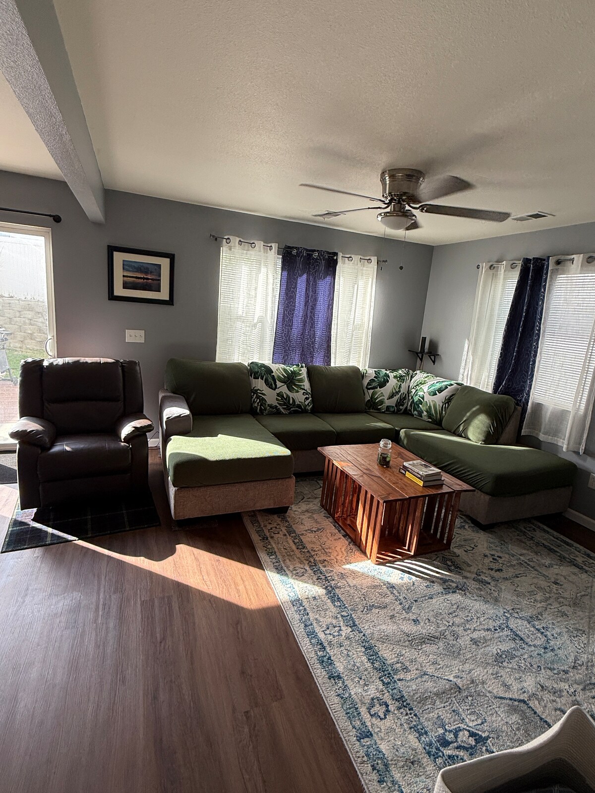 A well-lit living area features a large sectional sofa with green and floral accents. A wooden coffee table sits in front of the sofa. Natural light enters through two windows adorned with sheer curtains, creating a cozy atmosphere.