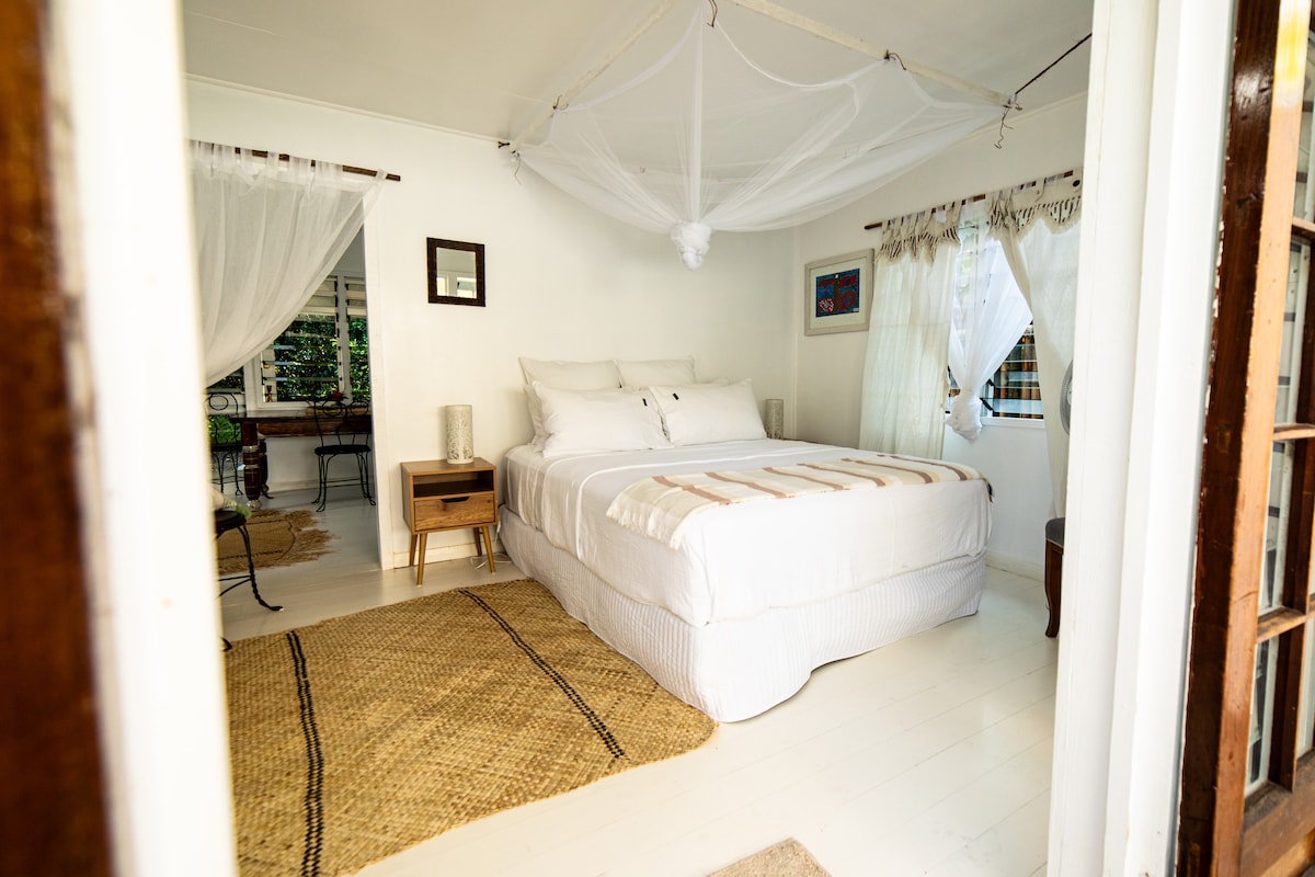 An inviting sleeping area features a spacious bed adorned with neutral linens and decorative pillows. Light flows through sheer curtains, while a woven rug adds a natural touch to the space. A bedside table and a chair complement the minimalist decor.