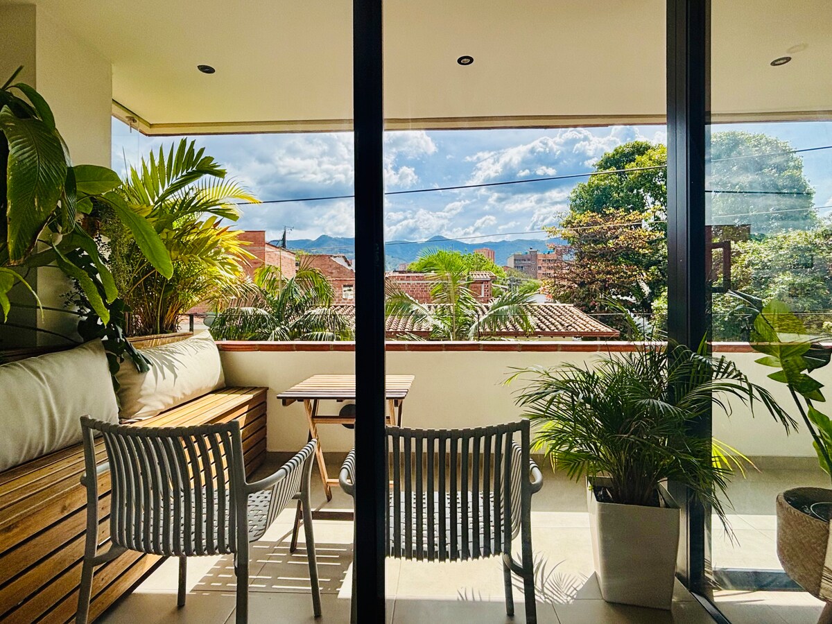 A spacious balcony is featured, furnished with a wooden bench and two white chairs. Lush green plants surround the area, creating a serene view of the city and mountains in the distance, while large glass doors allow natural light to fill the space.