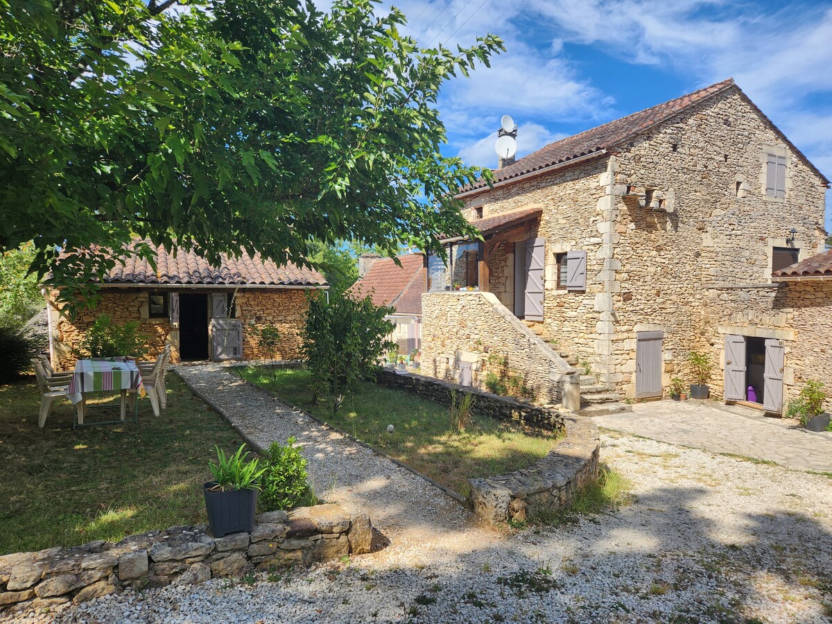 A stone house is depicted, surrounded by greenery and a gravel pathway leading to the entrance. Outdoor seating is visible on the left, accompanied by a shaded table. The sky shows a mix of blue and wispy clouds, enhancing the tranquil setting.