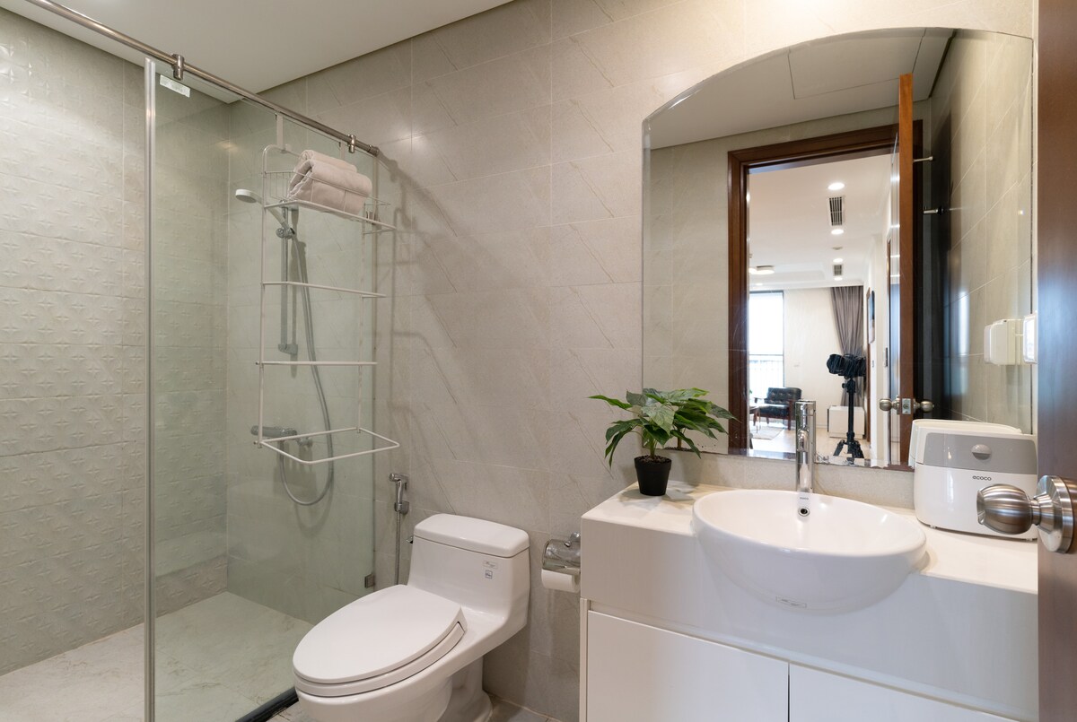 A modern bathroom is equipped with a glass shower enclosure and a sleek white toilet. A circular basin is mounted on the countertop, with a small plant adding a touch of greenery. Soft lighting enhances the clean lines and neutral tones.