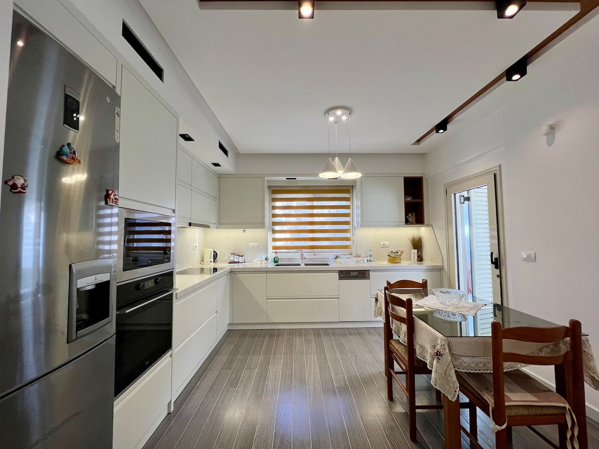 The modern kitchen is equipped with sleek cabinetry and stainless steel appliances, including a refrigerator and oven. A dining table with four wooden chairs is positioned next to a window with horizontal blinds, allowing for natural light to illuminate the space.