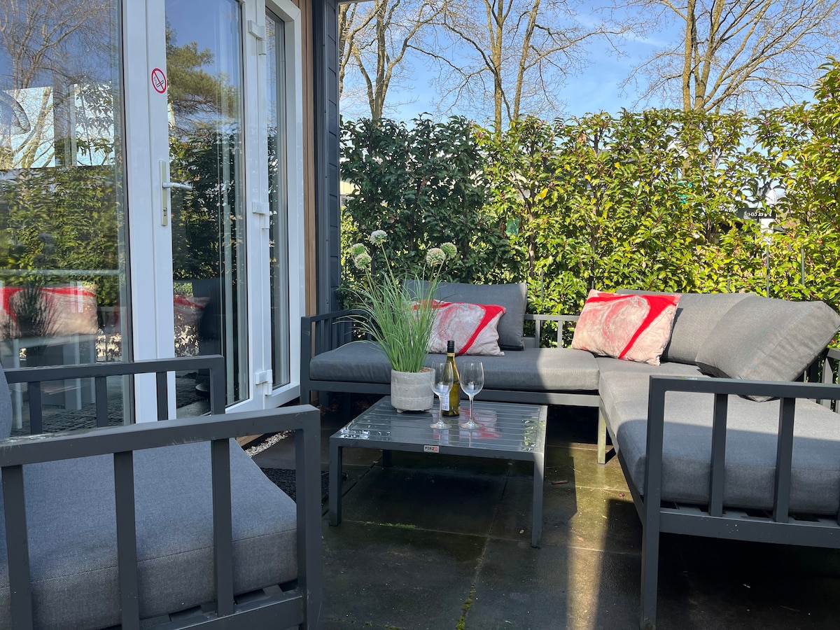 An outdoor lounge area is presented with gray seating arranged around a glass coffee table. Decorative plants are placed on the table, and a bottle of wine with glasses is visible, creating an inviting space. Surrounding greenery provides privacy.