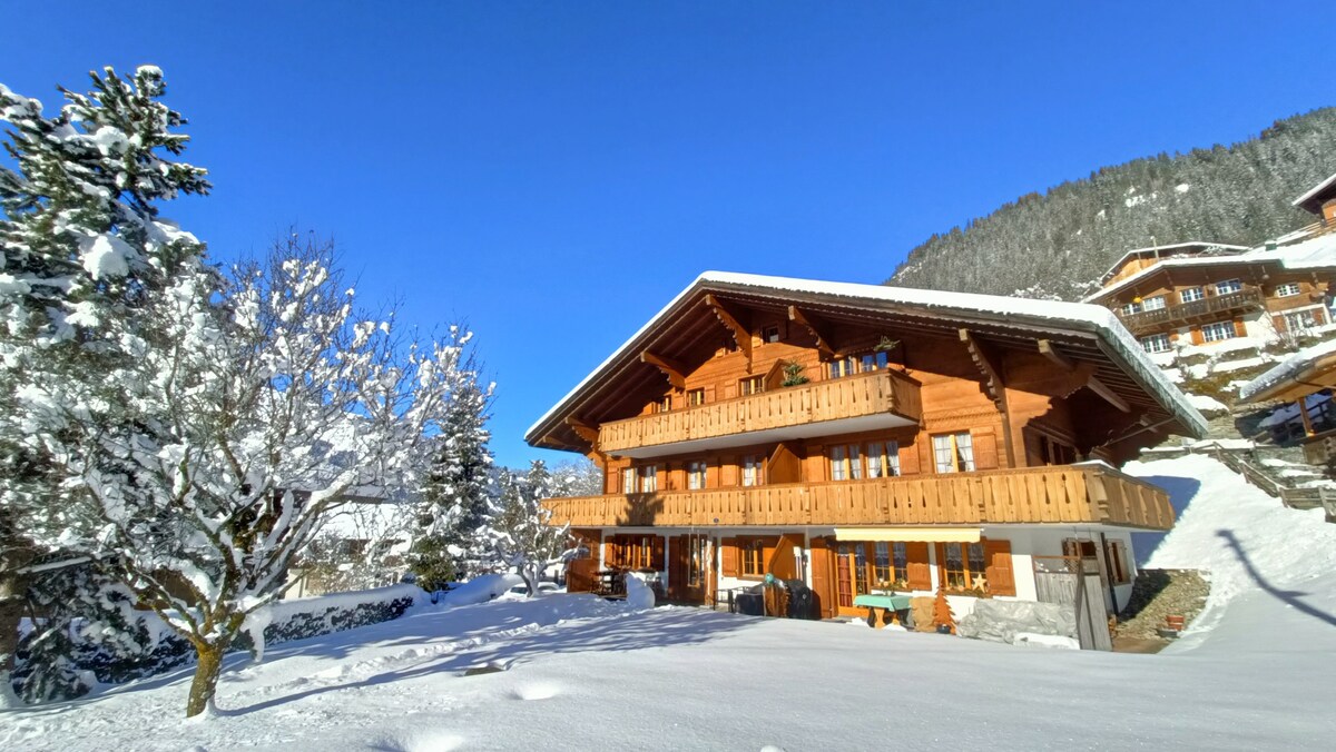 The charming chalet is surrounded by a pristine blanket of snow, showcasing its wooden balconies and detailed architecture against a clear blue sky. Snow-covered trees enhance the serene landscape, with the mountains rising gently in the background.