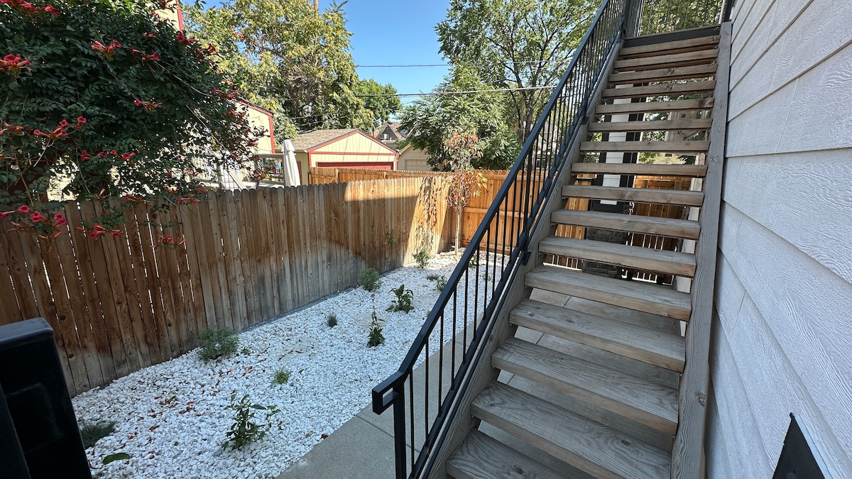 A wooden staircase is shown leading up to the entrance of the unit, surrounded by a gravel yard featuring sparse greenery. A wooden fence encloses the space, and several trees provide shade on a clear, sunny day.