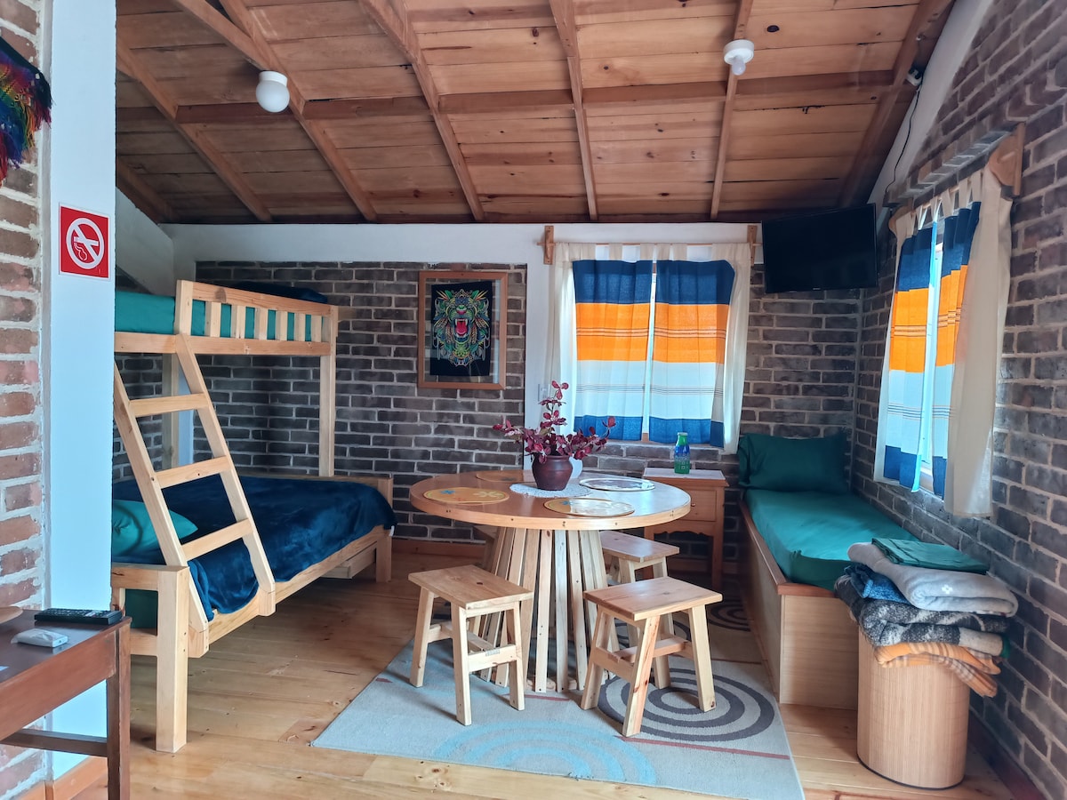 A cozy interior space features a wooden ceiling and exposed brick walls. A bunk bed occupies one corner, while a round dining table is accompanied by three chairs. A seating area with cushions is placed next to a window, adorned with colorful curtains that allow natural light in.