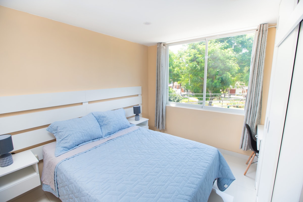 A well-lit bedroom features a spacious bed with soft blue bedding. Two bedside tables and lamps are positioned on either side, while a window offers a view of greenery outside. A small desk sits in one corner, providing an additional workspace.