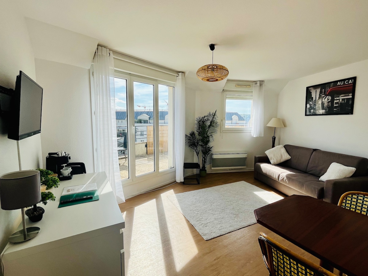 A spacious living area features large windows allowing natural light to fill the room. A comfortable sofa sits alongside a dining table, while a decorative plant adds a touch of greenery. Light-colored flooring complements the neutral color scheme of the furnishings.