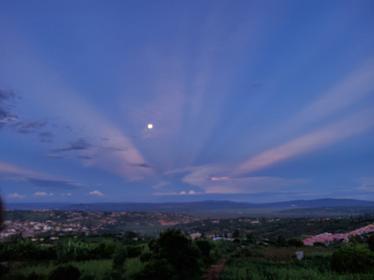 A scenic night sky features a full moon surrounded by soft, sweeping clouds. The horizon displays a landscape of hills, illuminated by the fading daylight. A tranquil atmosphere is conveyed through the subtle hues of blue and pink that stretch across the sky.