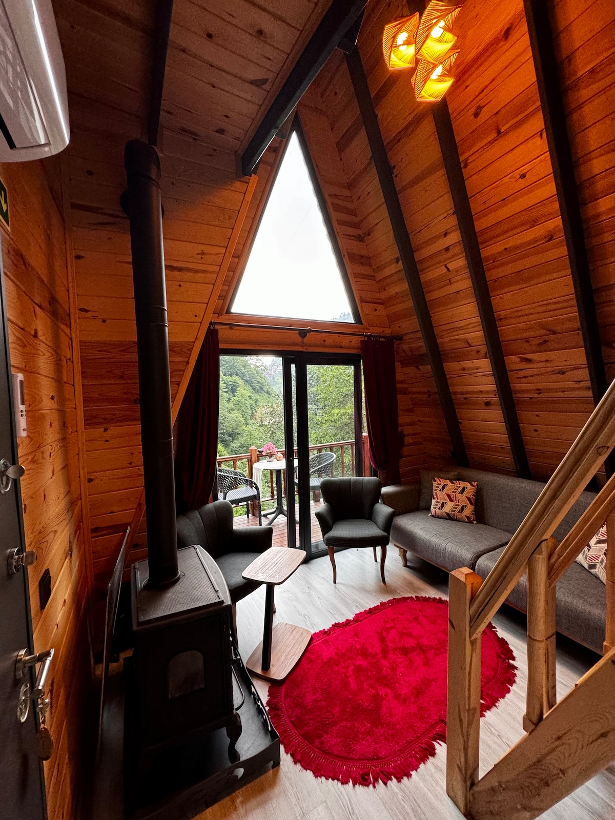 An inviting interior features a triangular wooden structure with large windows allowing natural light. A cozy seating area includes a gray sofa and two small side tables. A red area rug adds a pop of color, while a wood-burning stove enhances the warmth of the space.