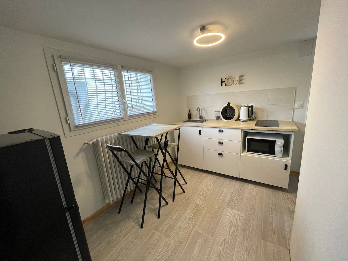 A compact kitchen area features a minimalist design with white cabinetry and a small dining table paired with two stools. Appliances, including a microwave and coffee maker, are arranged for convenience. Natural light enters through the partially opened window with white blinds.