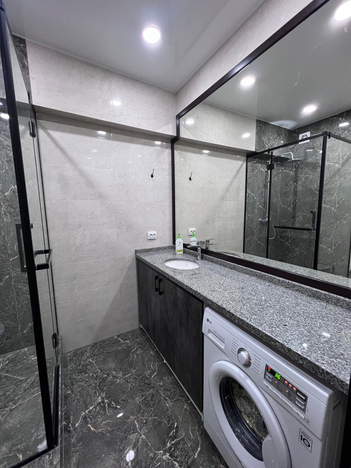 A modern bathroom features sleek grey marble walls and flooring. A glass shower enclosure is present, alongside a contemporary washing machine. A large mirror above a granite countertop reflects the well-lit space, which is equipped with minimalist fixtures for convenience.
