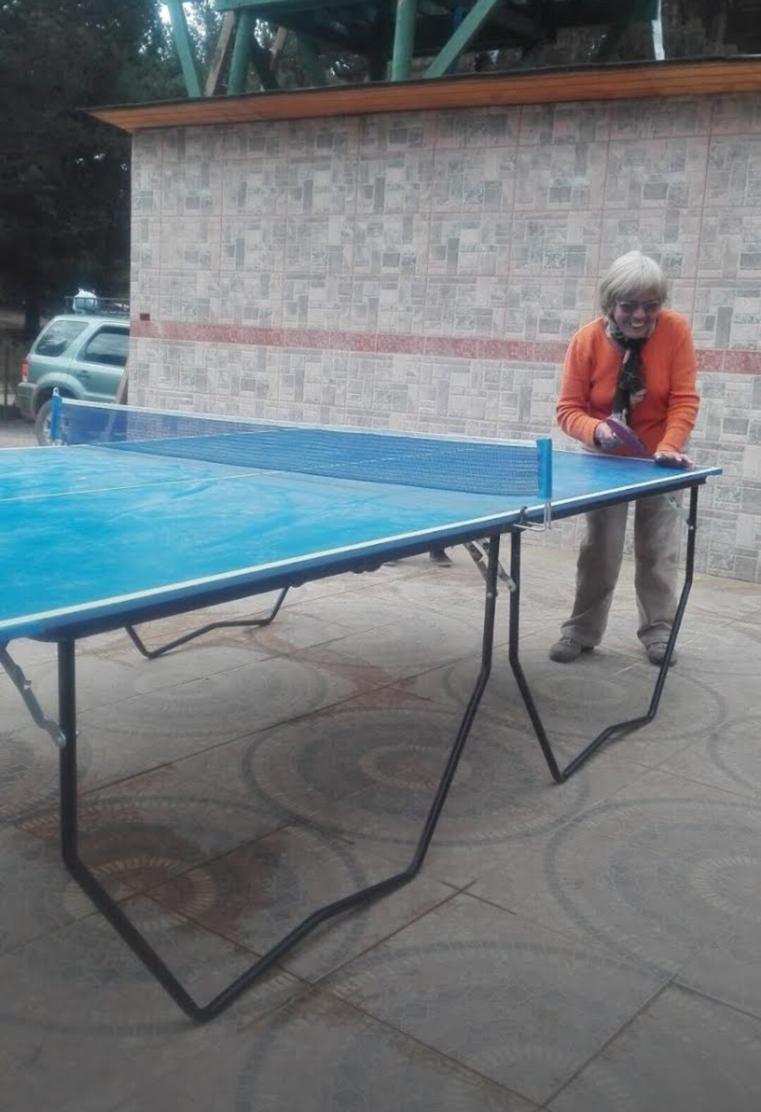 A blue ping pong table is set up outdoors on a tiled surface, featuring sturdy black legs. A person is seen adjusting the table, while a vehicle is parked in the background near a textured wall.