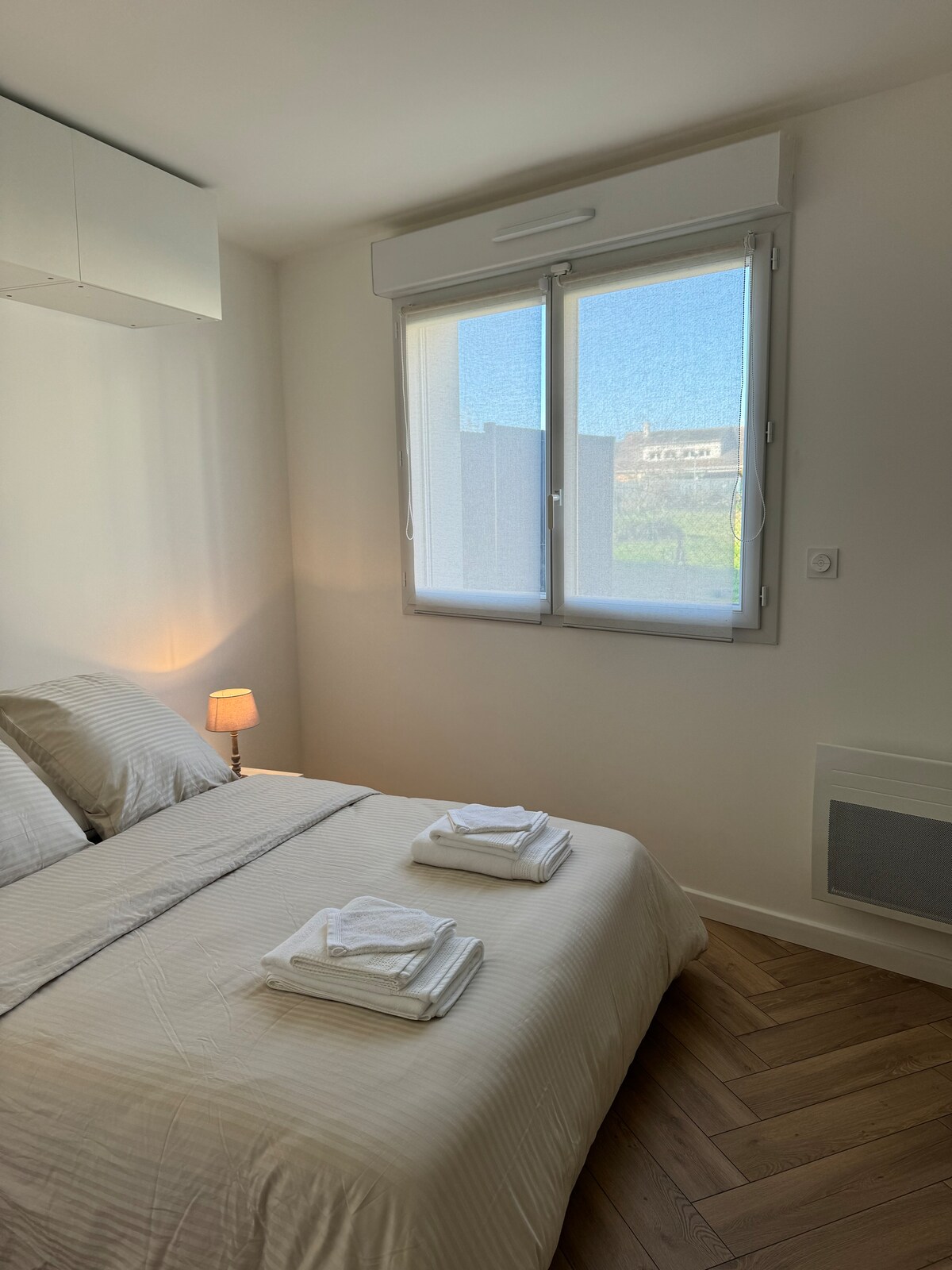 A well-lit bedroom features a spacious bed with neatly arranged towels on top. Natural light enters through a window with sheer coverings, illuminating the minimalist decor. A small lamp beside the bed adds a touch of warmth to the tranquil atmosphere.