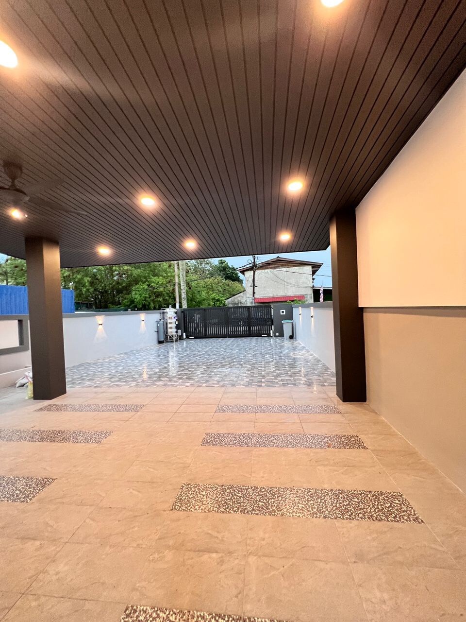 An outdoor area characterized by a smooth tiled floor and well-placed lighting fixtures. The space is bordered by a clean, low wall, providing a sense of openness while maintaining privacy. The entrance features a secured gate, ensuring a welcoming and secure environment.