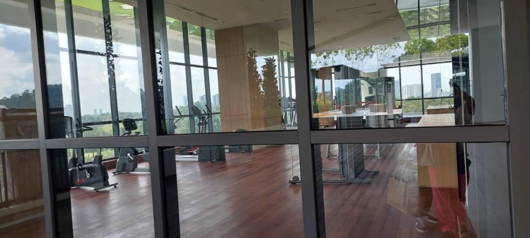 A spacious fitness area is visible through large glass doors, featuring wooden flooring and floor-to-ceiling windows. Modern exercise equipment is arranged in the background, with natural light illuminating the space. Views of greenery can be seen outside, creating a calming environment.