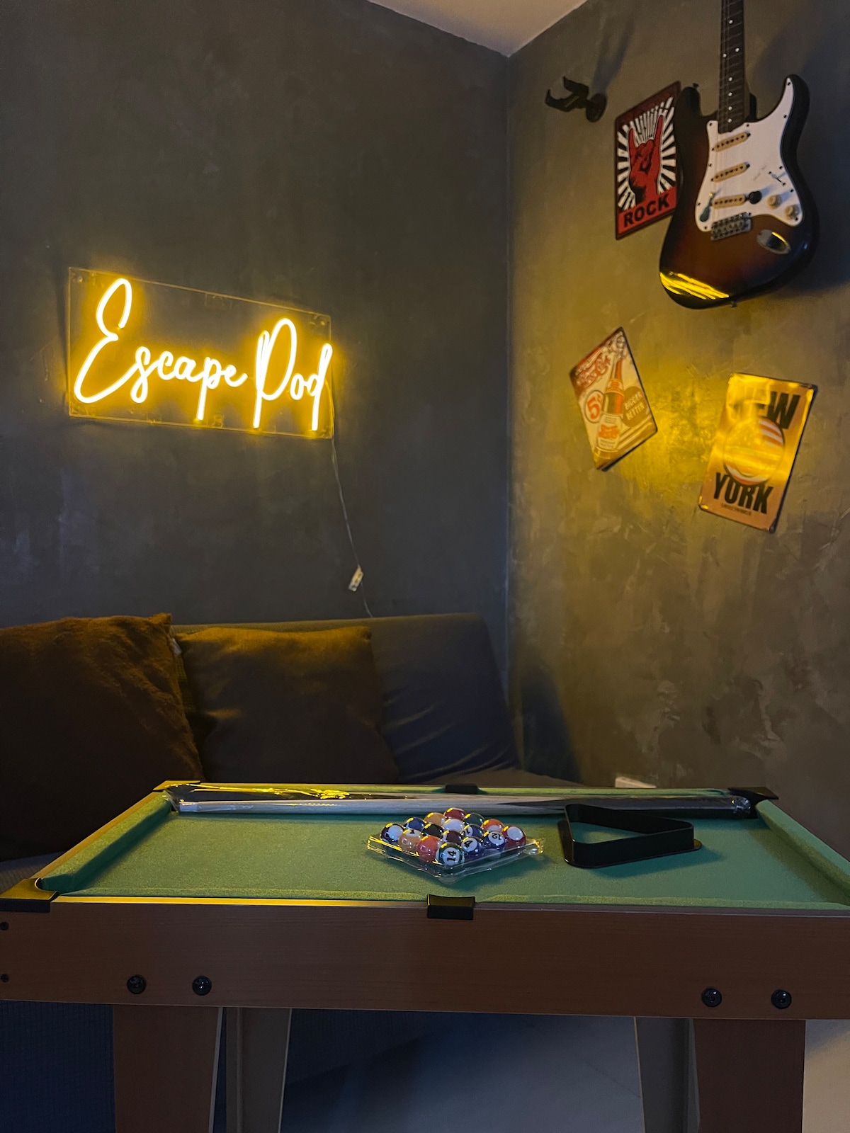 A cozy corner is showcased with a comfortable couch in muted tones and a vibrant neon sign reading 'Escape Pod.' A small billiard table is set up in front, complete with colorful balls. Rock-themed artwork adorns the walls, enhancing the playful ambiance.