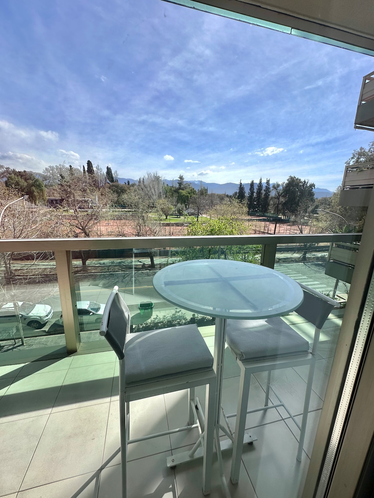 A balcony features a circular glass table with two high-backed chairs, overlooking a view of greenery and distant mountains. A bright sky is visible, enhancing the outdoor setting.