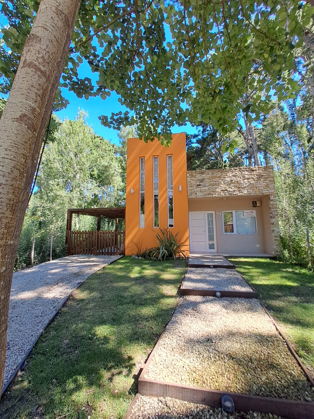 A modern home features an orange exterior with large vertical windows, surrounded by greenery. A gravel pathway leads to the entrance, with a covered carport visible beside the house, providing a welcoming approach in a tranquil forest setting.