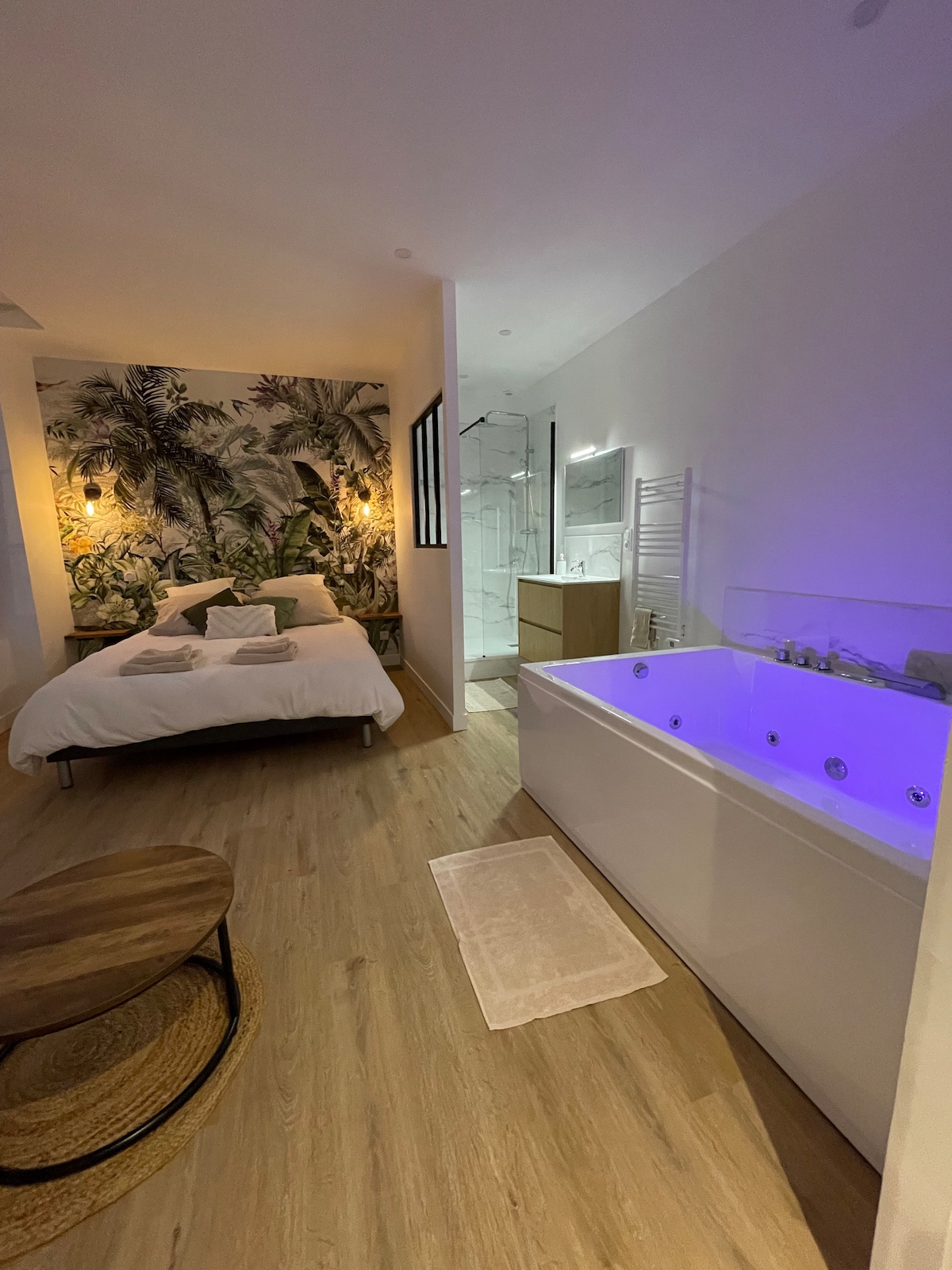 A modern bedroom features a spacious bed adorned with greenery-inspired wall art. A chic, illuminated whirlpool bathtub is visible in the corner, accompanied by a wooden coffee table and a soft area rug on the light wood floor.