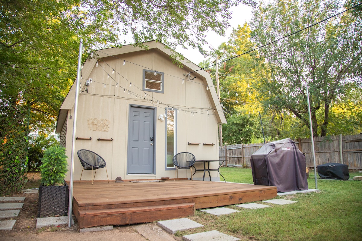 A charming outdoor area features a wooden deck leading to a light-colored house. String lights add ambiance, while two black chairs are placed near a small table. A grill is located to the side, surrounded by green grass and mature trees.