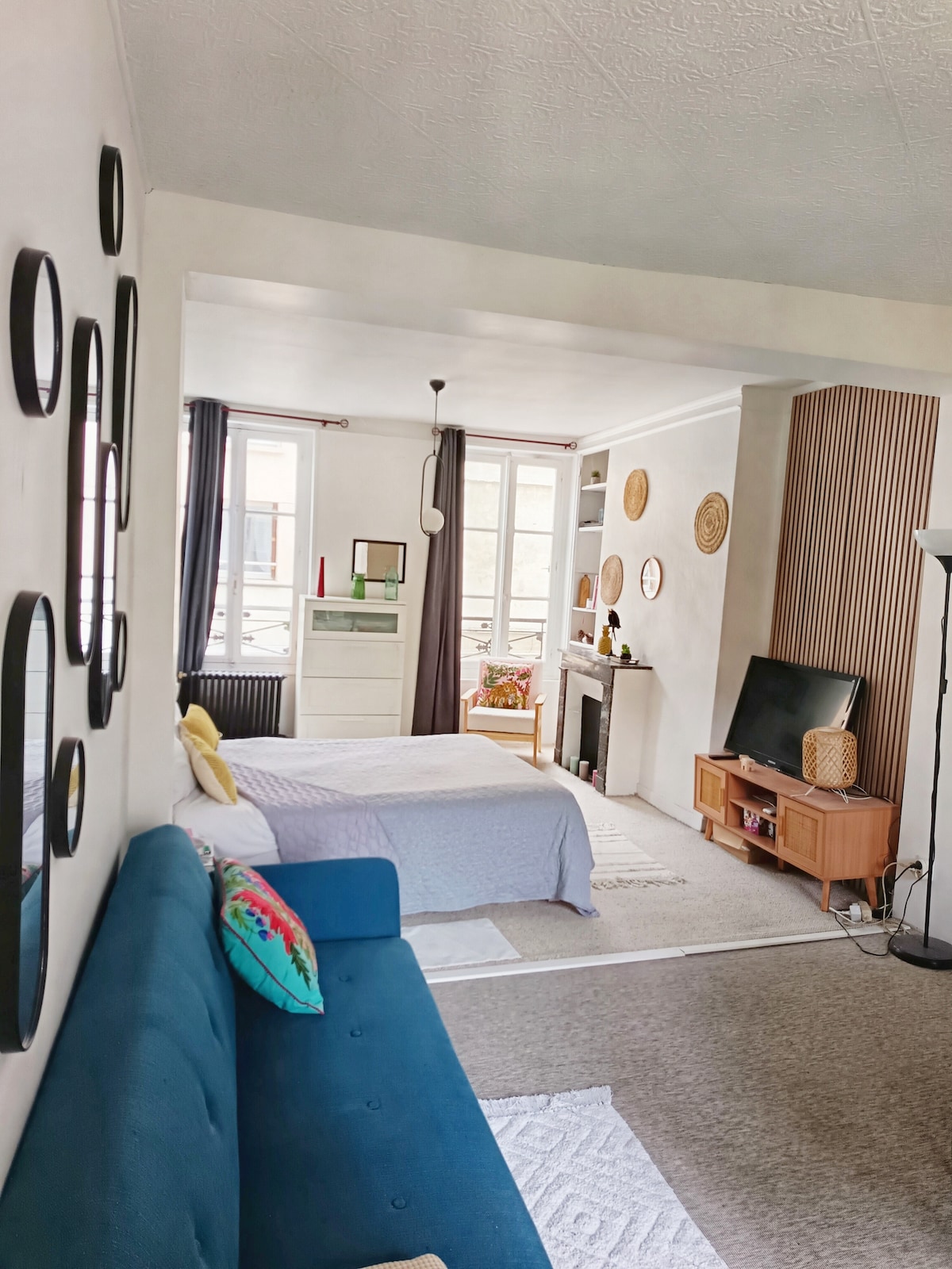 A comfortable living space features a spacious bed and a cozy sofa, complemented by decorative mirrors on the walls. Natural light brightens the area through large windows, with neutral tones enhanced by colorful cushions and wall accents. A television and decorative fireplace add to the ambiance.