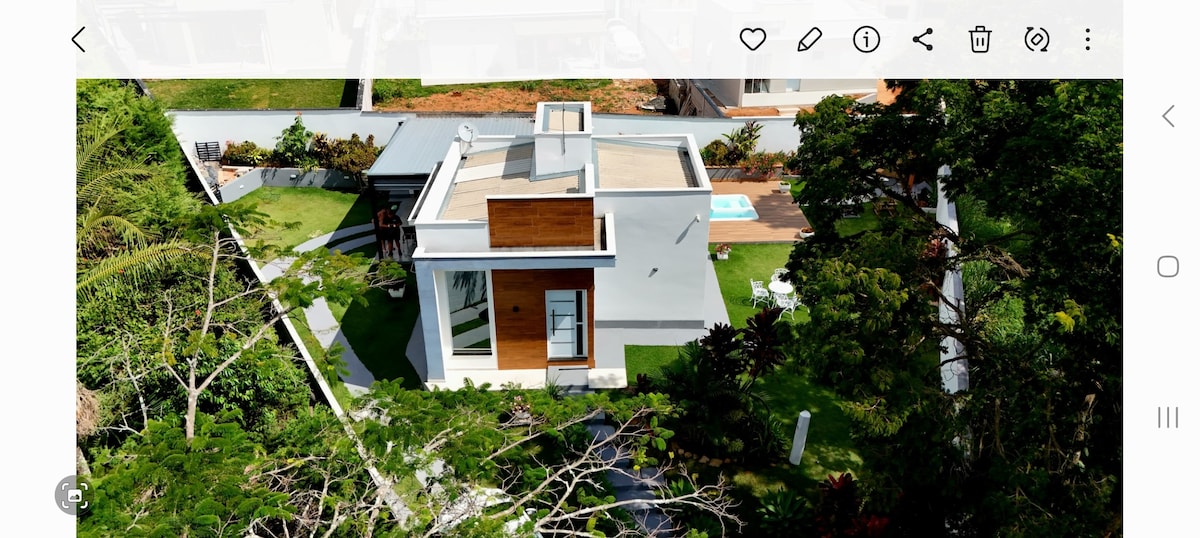 An aerial view of a modern house is captured, showcasing a flat roof and large windows. Surrounding greenery and a well-kept lawn enhance the outdoor space, with a clear pool visible in the background. A shaded area features outdoor seating.