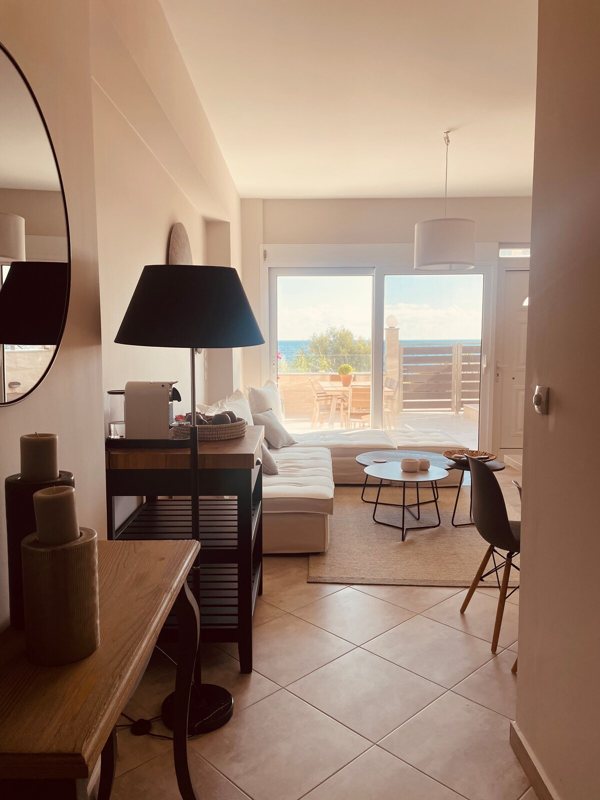 A bright entryway leads to a lounge area, featuring a comfortable white sofa and a round coffee table. Large glass doors provide a view of the sea and open to an outdoor seating space. Natural light fills the room, enhancing the inviting atmosphere.