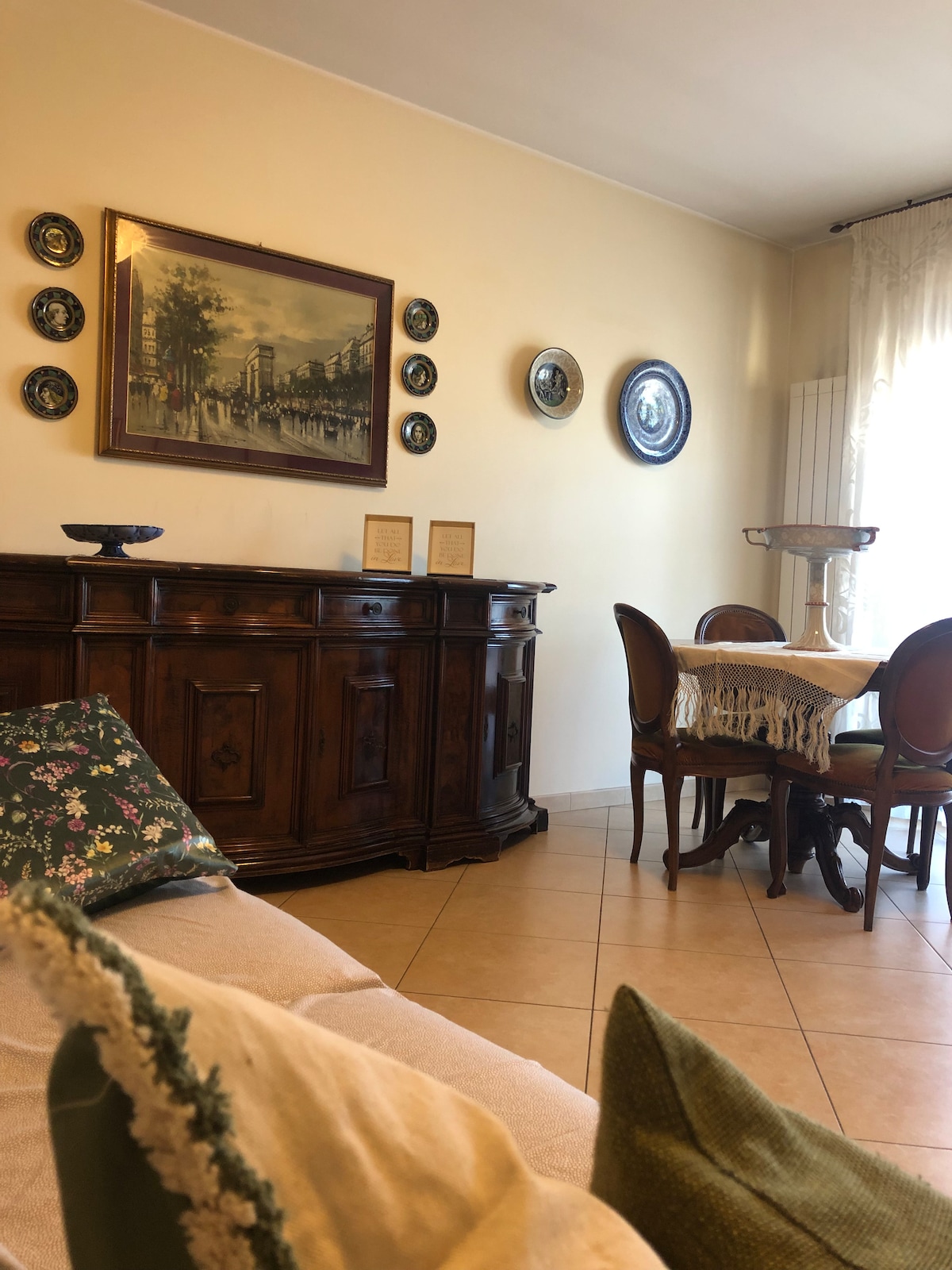A warm living area features a wooden sideboard adorned with decorative plates and artwork on the walls. A dining table with elegant chairs is positioned nearby, while light enters through the window, complemented by soft drapes. Cushions add comfort to the seating space.