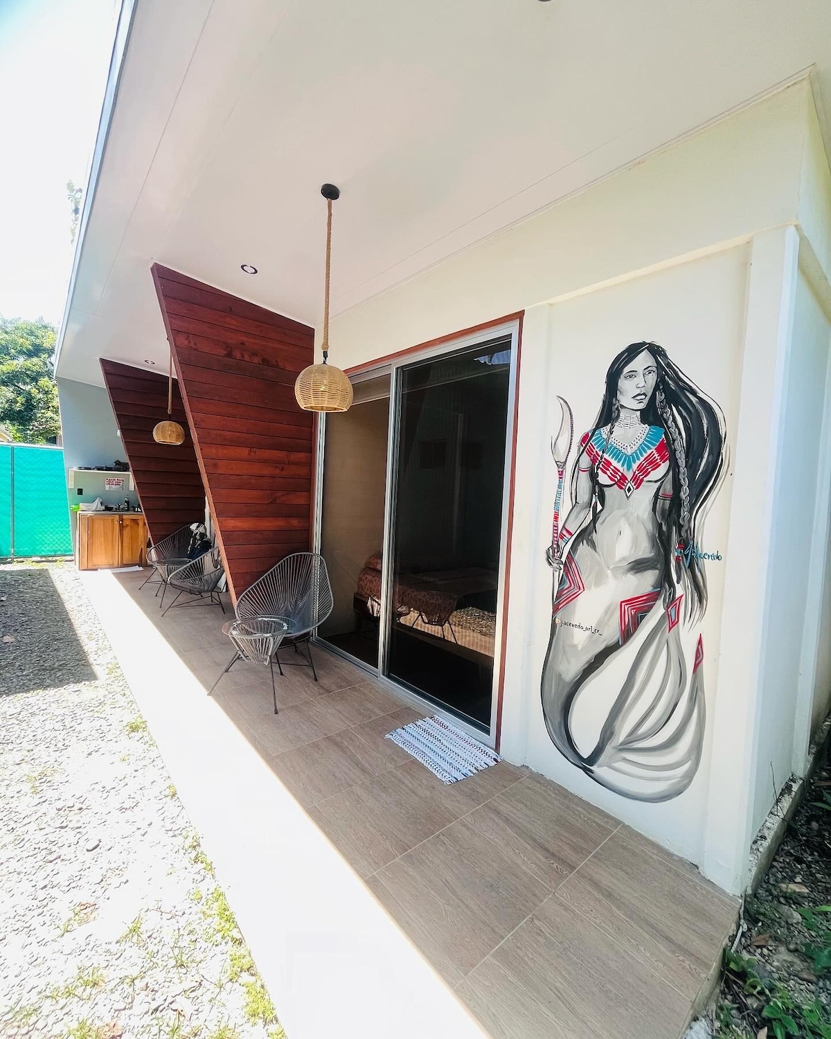 The exterior of the cabana-style accommodation features a striking mural of a figure adorned in vibrant jewelry. Two unique wooden structures create a distinctive architectural silhouette. A spacious patio area is accessible through large sliding doors, leading to an inviting outdoor environment.